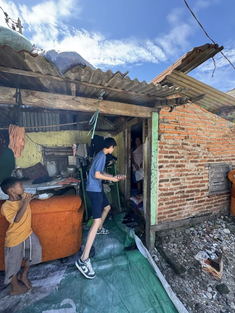 A volunteer steps into a home with a tin roof while a young boy looks on