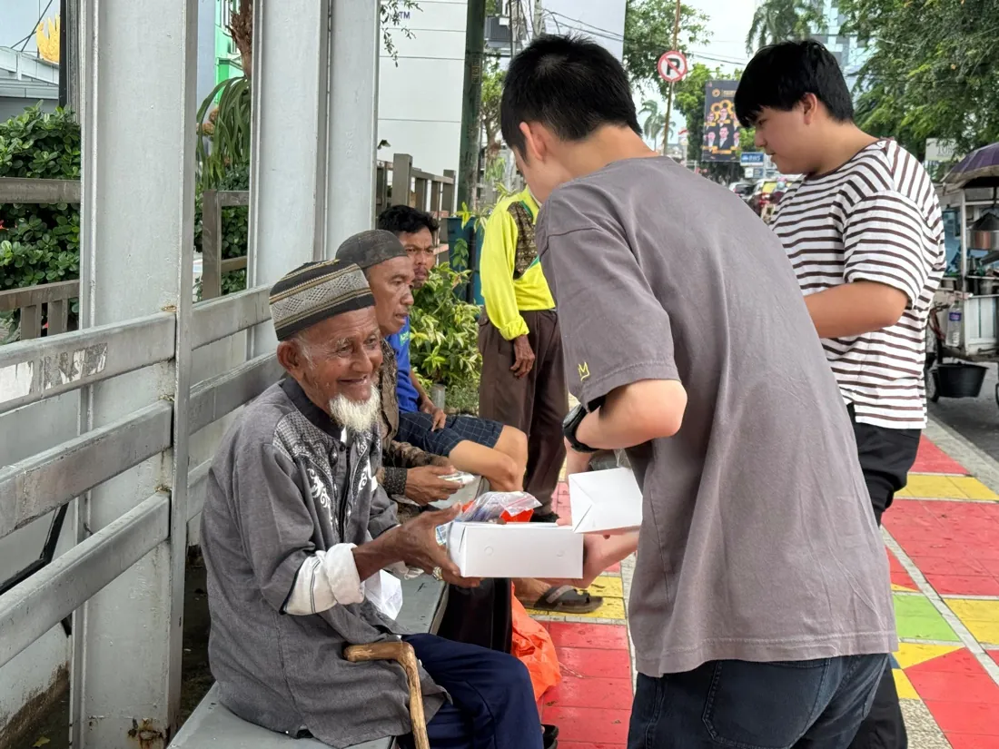 An older man on a bench smiles as volunteers hand him a care package